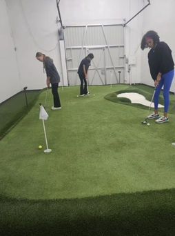 Three people golfing indoors on an artificial turf putting green, with clubs and a flag cup.