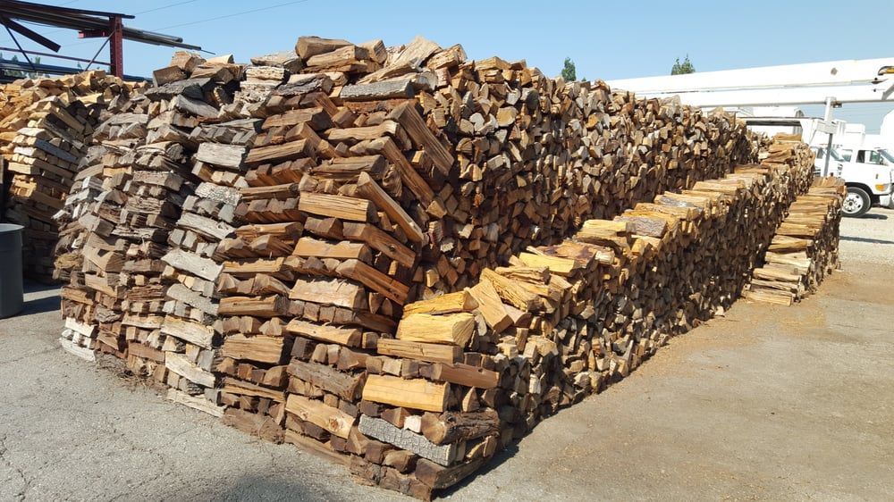 Stacks of cut firewood piled high outdoors in a yard, with a white building in the background