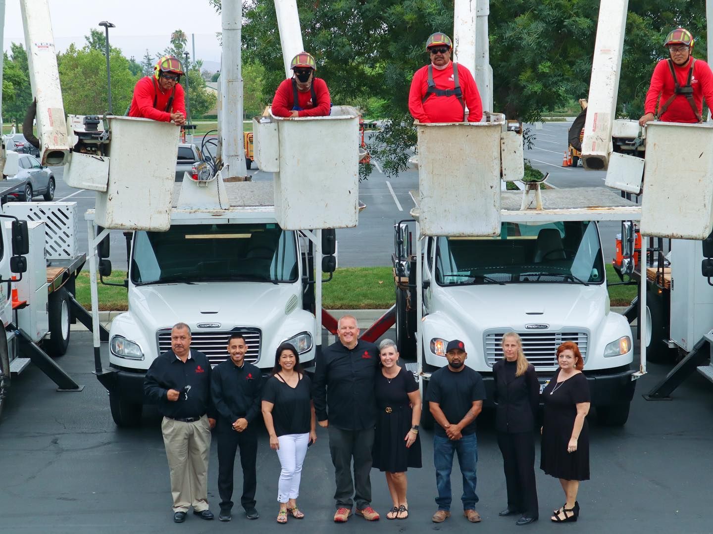 Group of workers posing in front of two white utility trucks with bucket lifts, red-clad operators above.