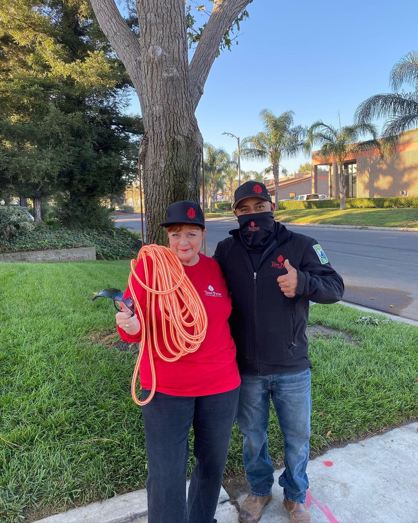 Two people outdoors by a tree and street, one holding orange rope and both giving thumbs up.