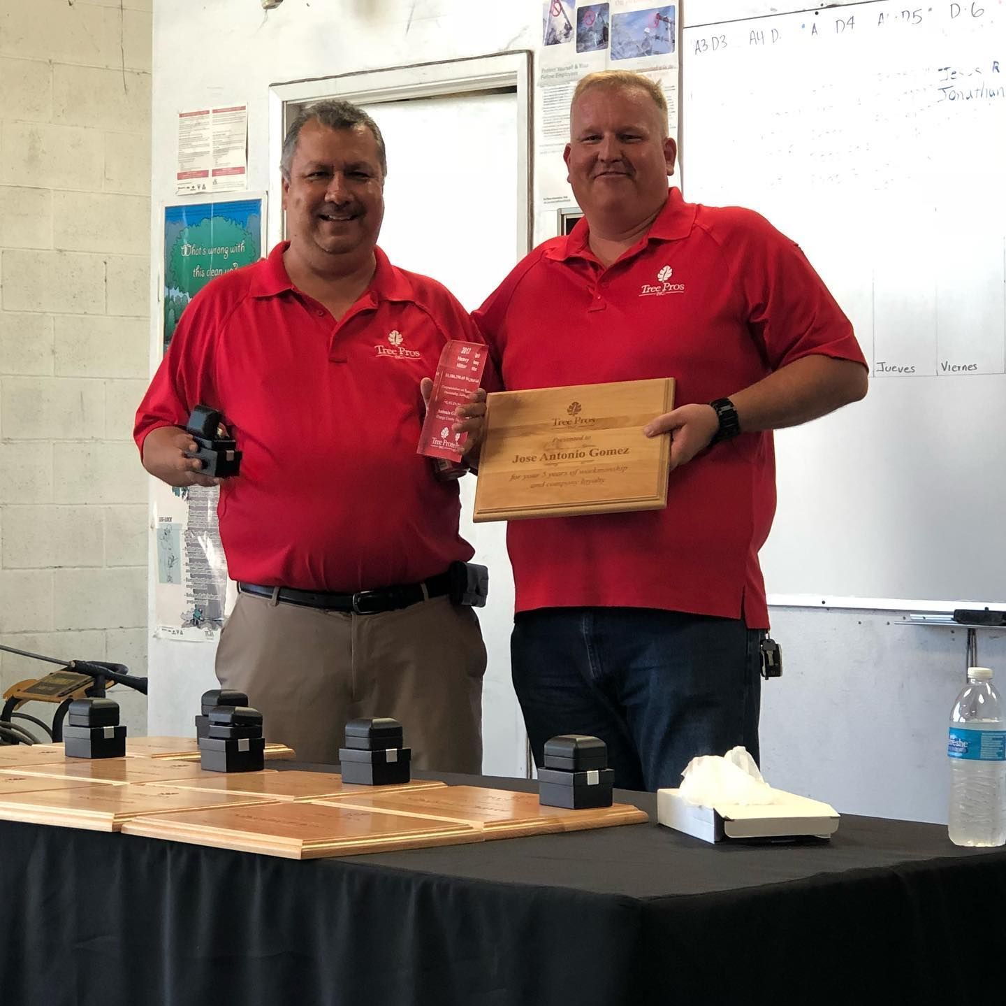 Two men in red shirts hold a plaque and drink in a classroom or office setting.