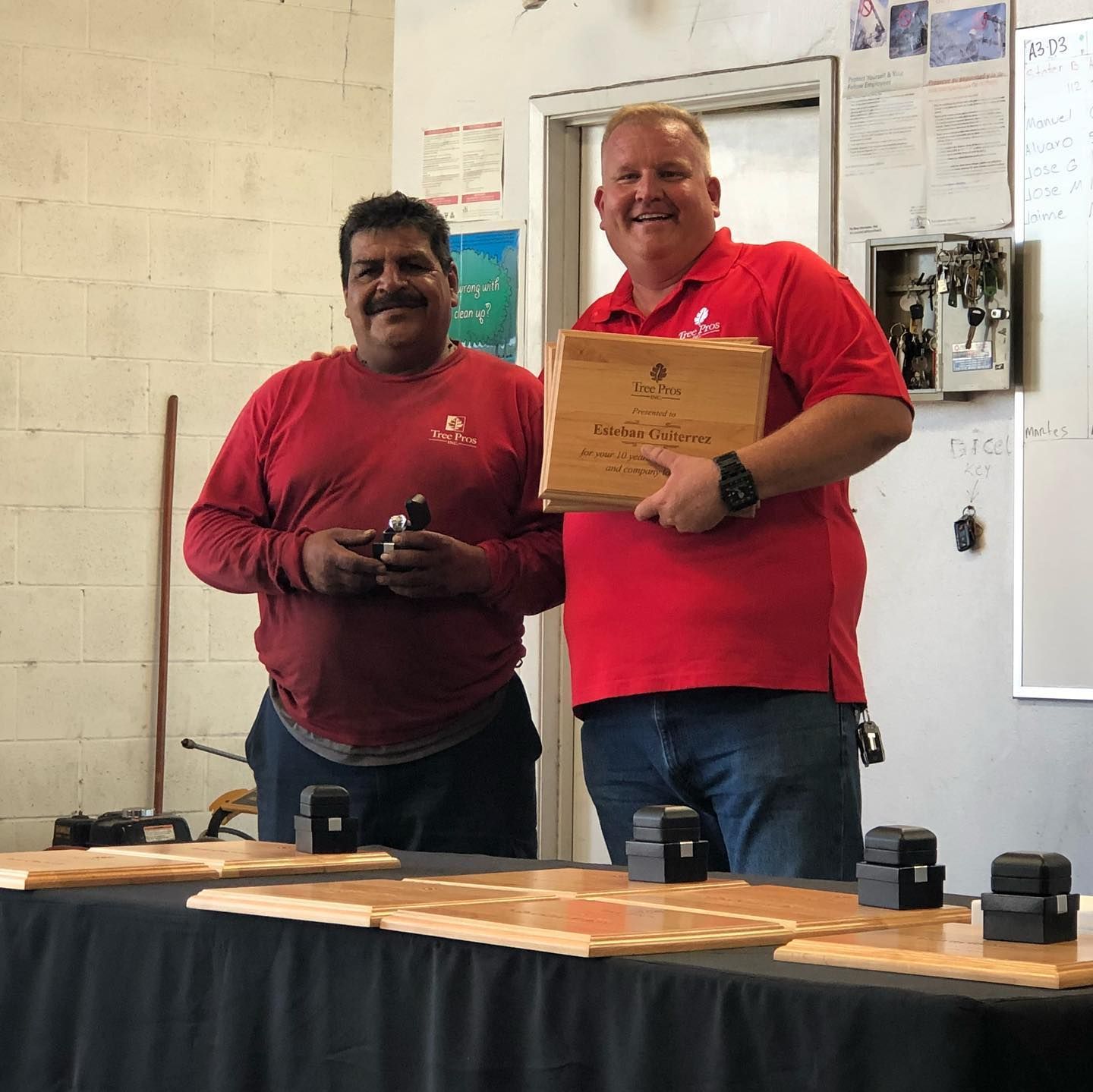 Two men in red shirts pose indoors; one holds a plaque behind a table with black boxes.