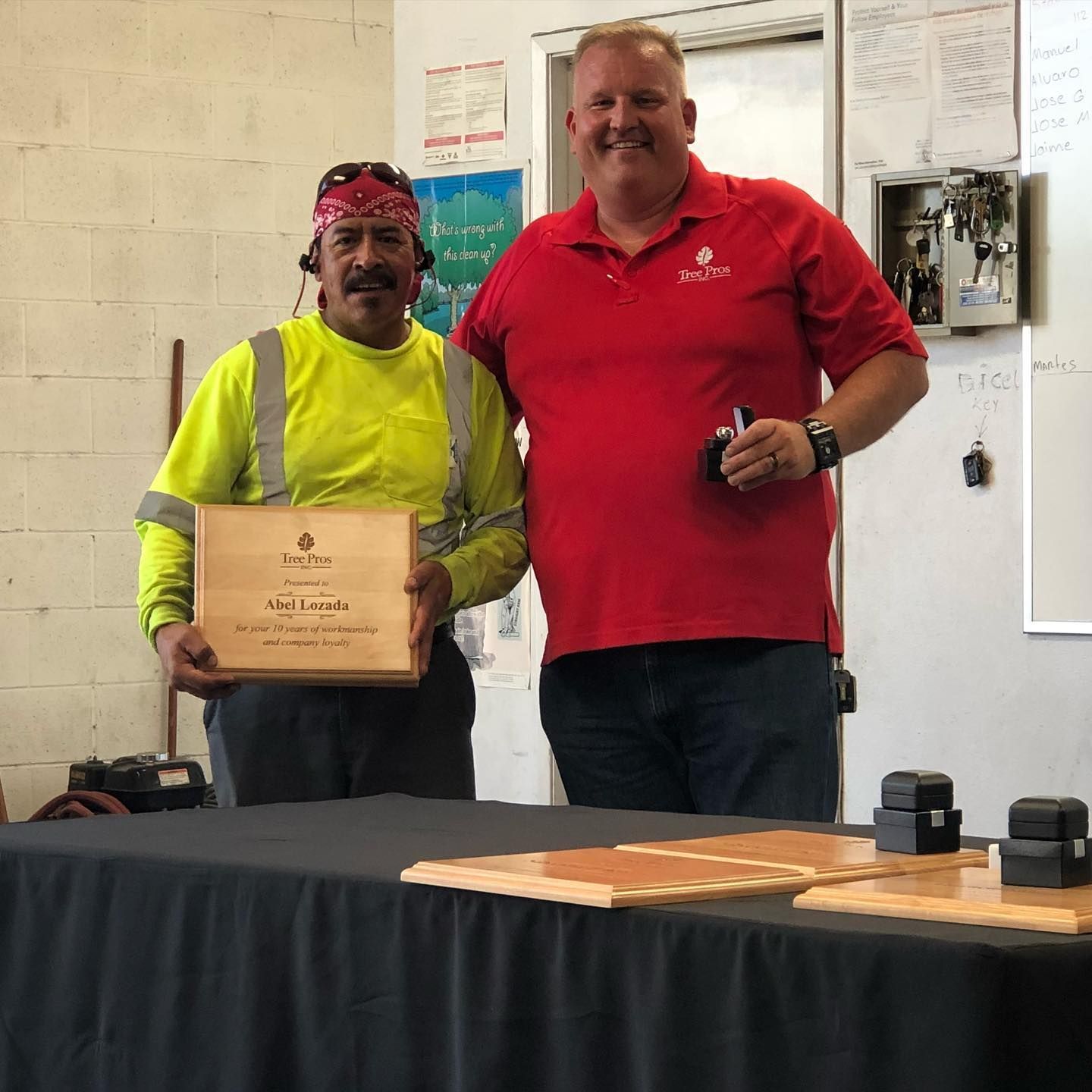 Two men stand behind a table, one holding a plaque and the other holding a small item, in a workshop or office.