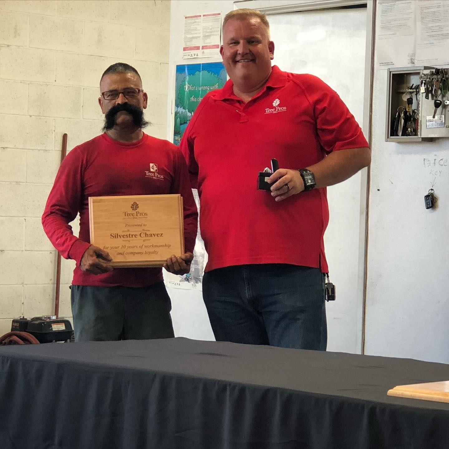 Two men in red shirts stand at a table, one holding a plaque, in an indoor award setting.
