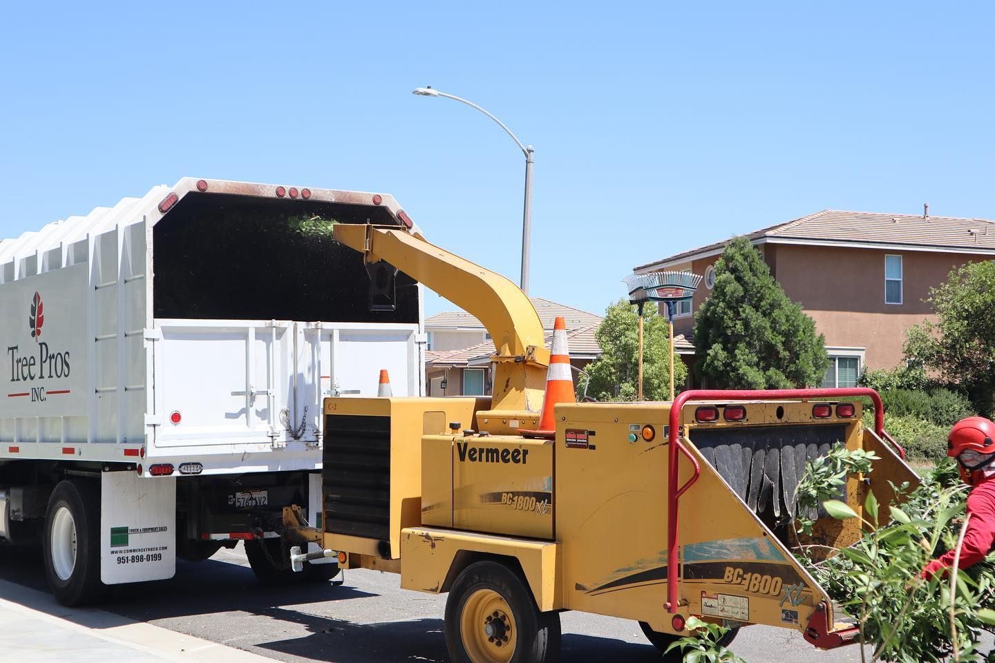 Yellow wood chipper blowing debris into a white truck on a sunny suburban street