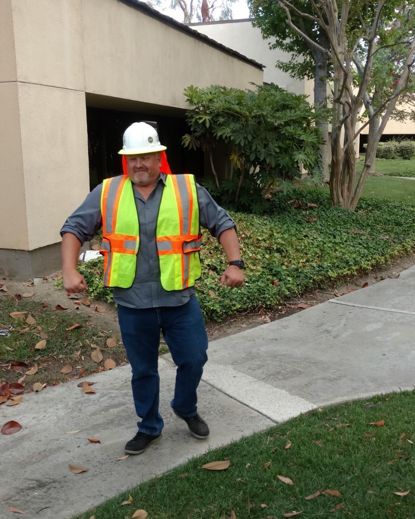 Construction worker in hard hat and safety vest standing on a sidewalk near a building.