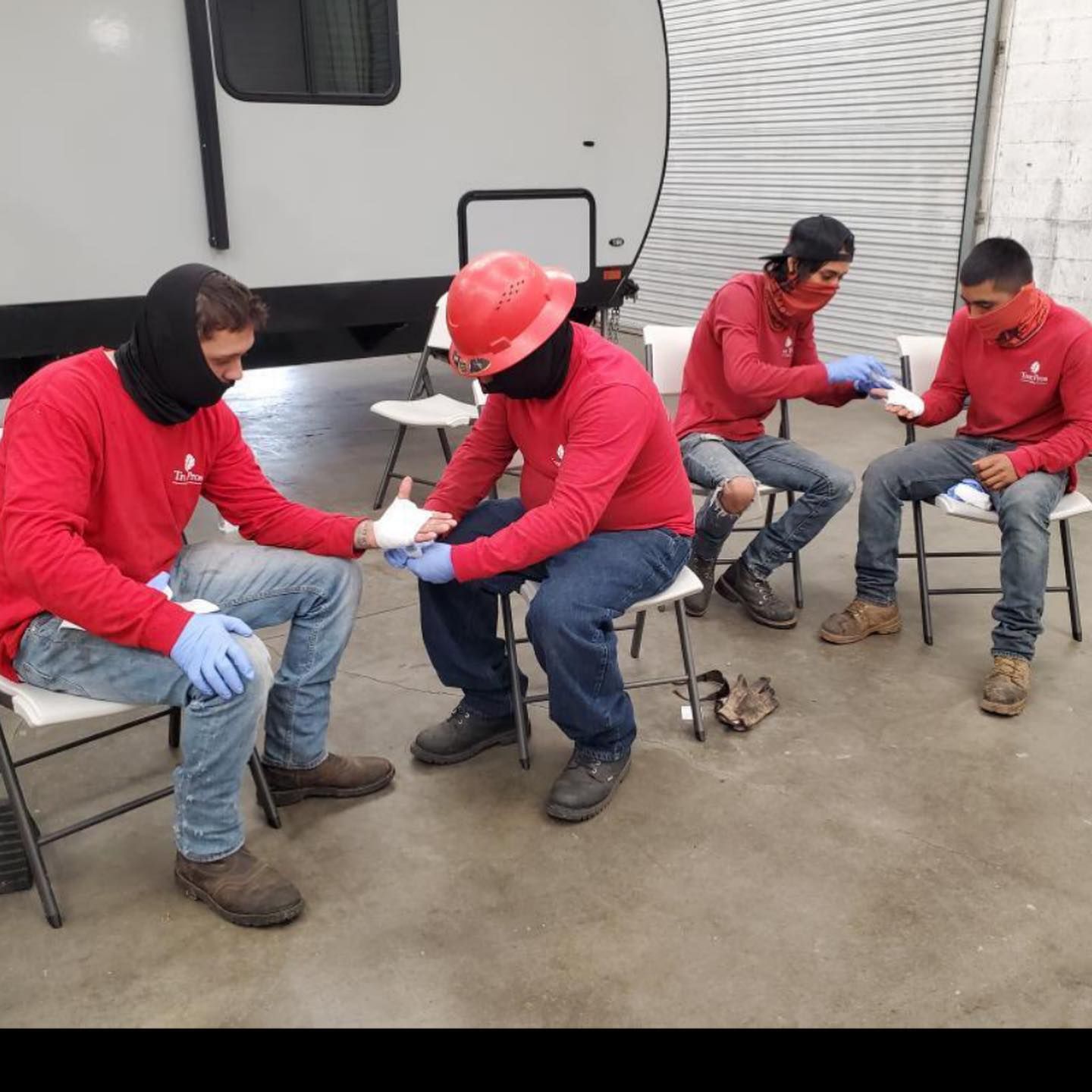 Four workers in red shirts and helmets sit on chairs in a warehouse, taking a break.