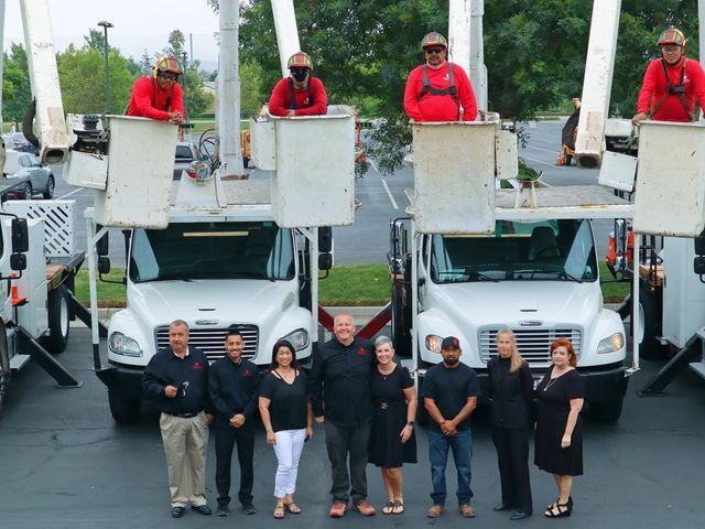Group posing in front of two utility trucks with workers on elevated platforms at a worksite