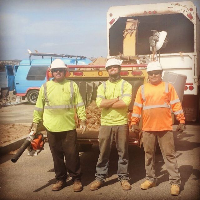 Three workers in hard hats and hi-vis vests stand beside construction equipment at a worksite.