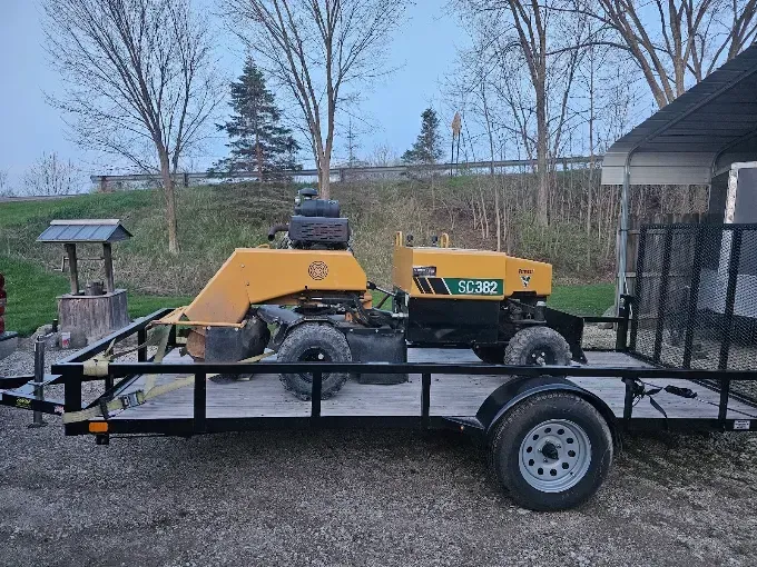 Yellow trailer-mounted machine parked outdoors near a building and trees