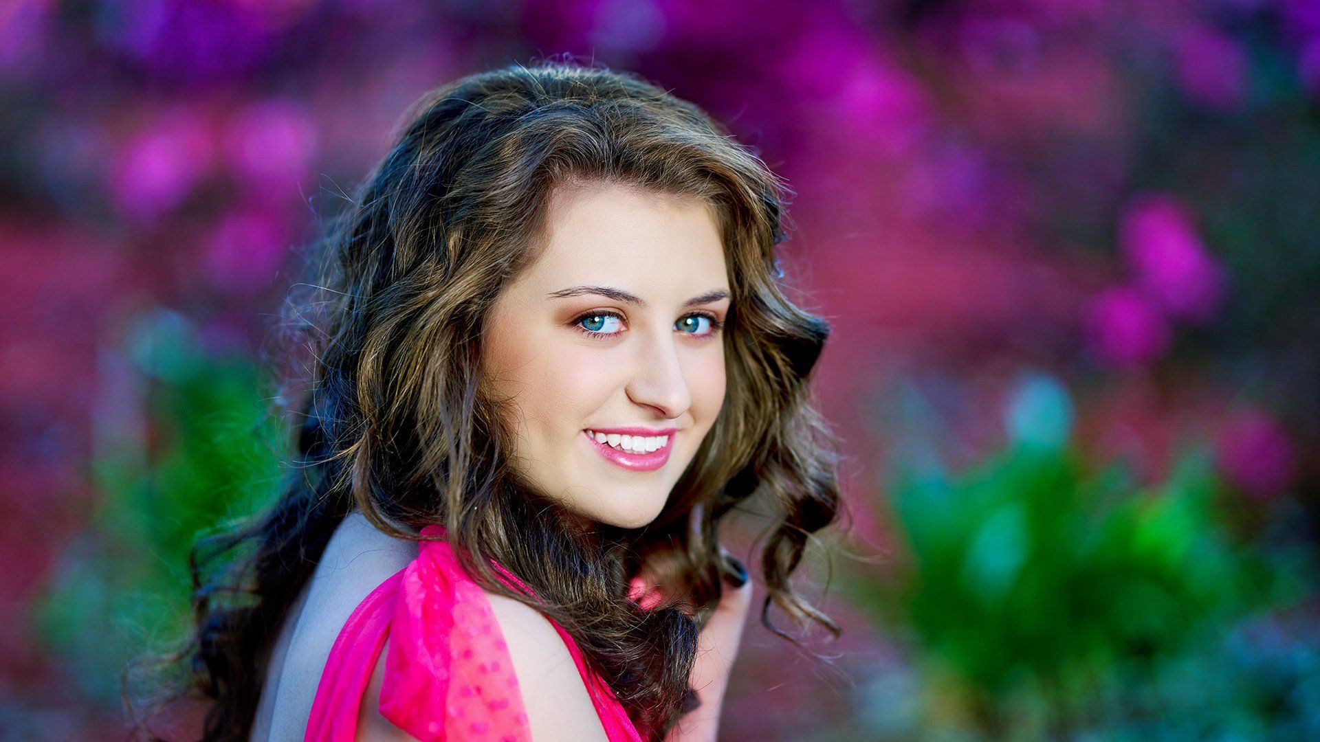 A woman in a pink dress is smiling in front of purple flowers.