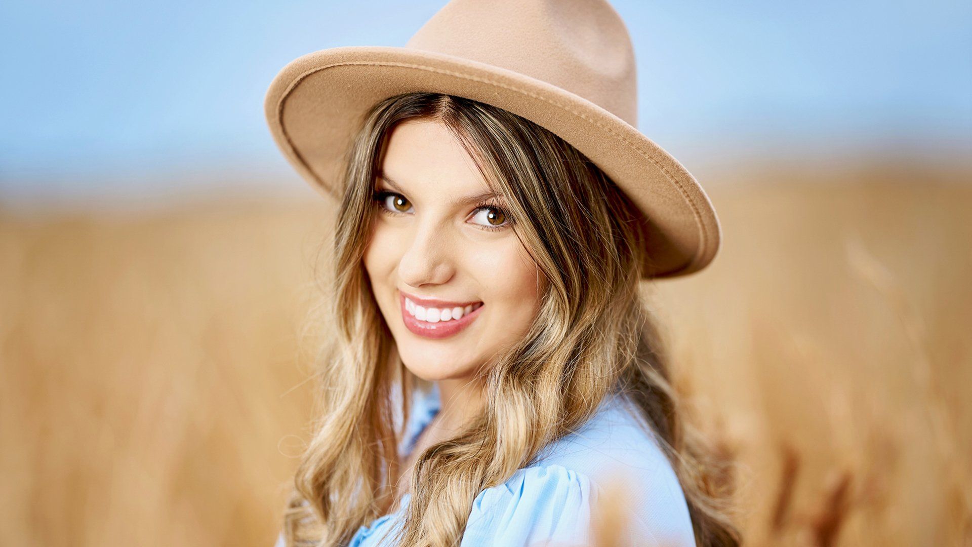 A woman wearing a hat and a blue shirt is standing in a field of wheat.