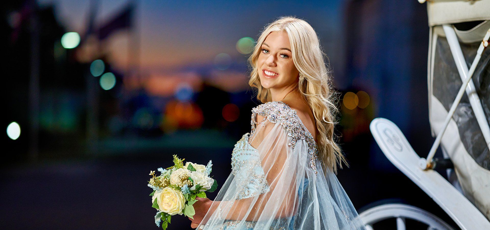 A woman in a blue dress is standing next to a white carriage holding a bouquet of flowers.