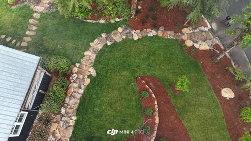 Aerial view of a landscaped garden with green lawn, red mulch beds, stepping stones, and a house roof edge