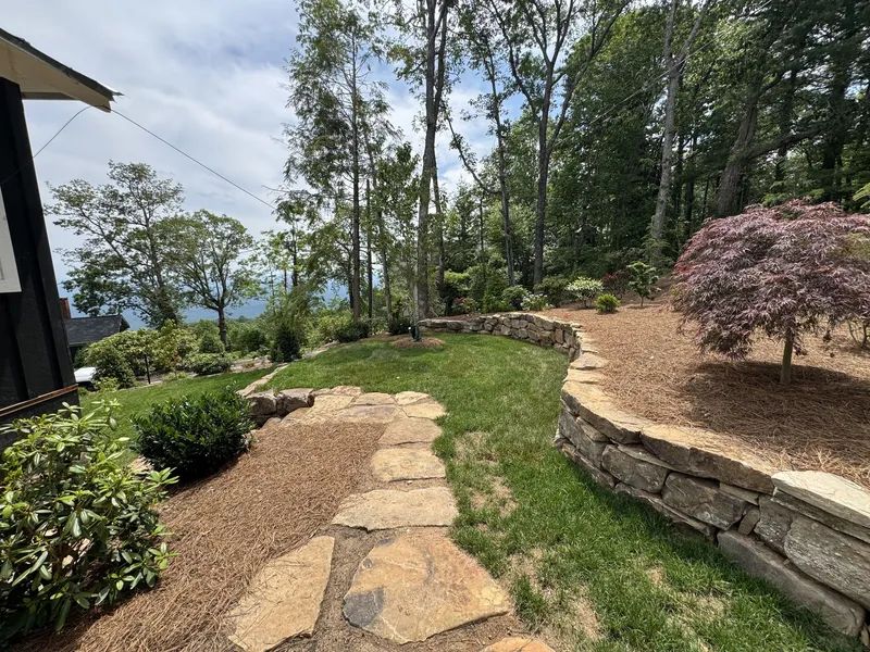 Stone path through a landscaped garden with trees, grass, and a low retaining wall on a sunny day