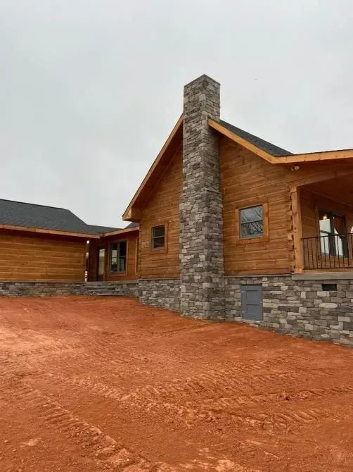 Log cabin-style house with stone chimney and red dirt driveway under overcast sky