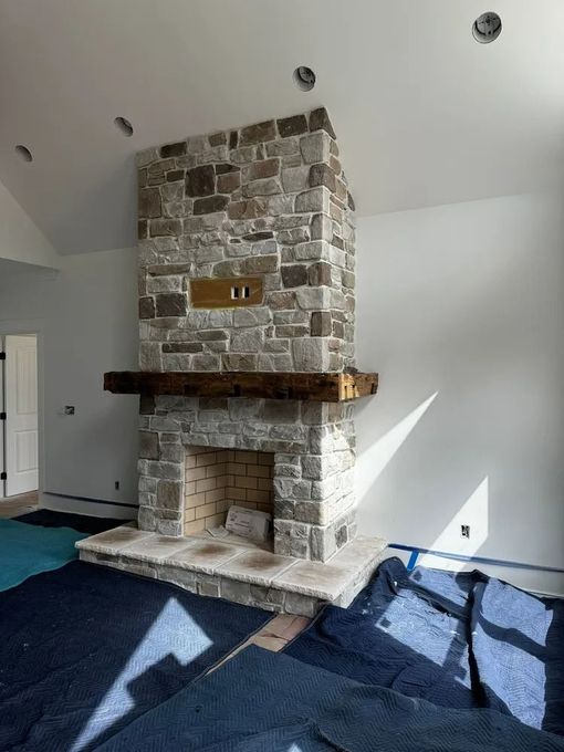 Stone fireplace with wood mantel in a bright living room with dark carpet and sunlight on the wall
