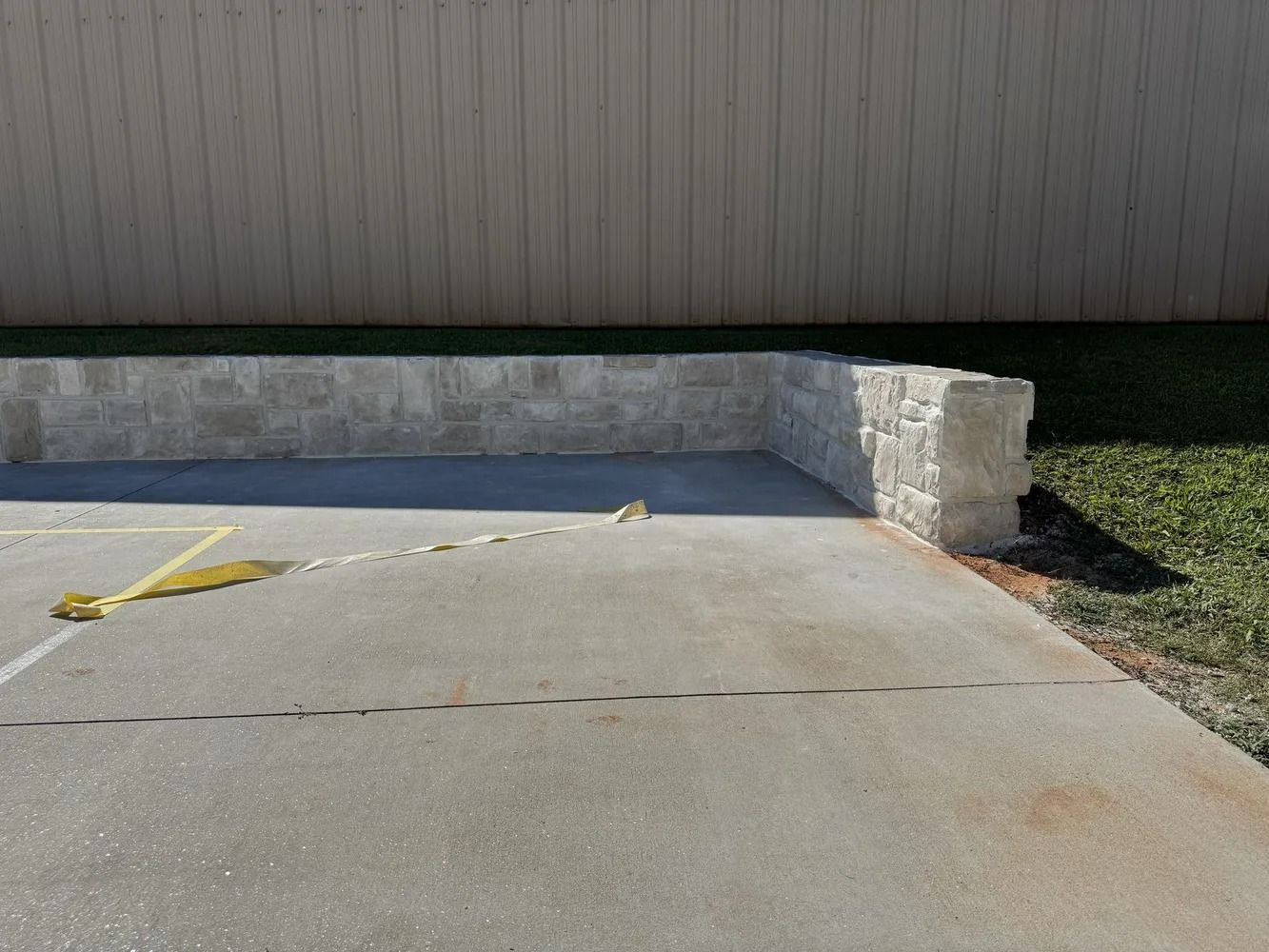 Concrete parking pad beside a stone retaining wall and beige building, with yellow wheel stops.