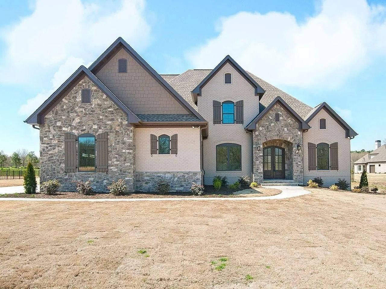 Stone house with gabled roof and blue shutters on a large bare front yard under a partly cloudy sky