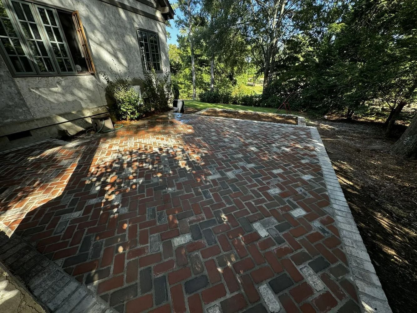 Brick patio beside a house, shaded by trees with dappled sunlight and fallen leaves