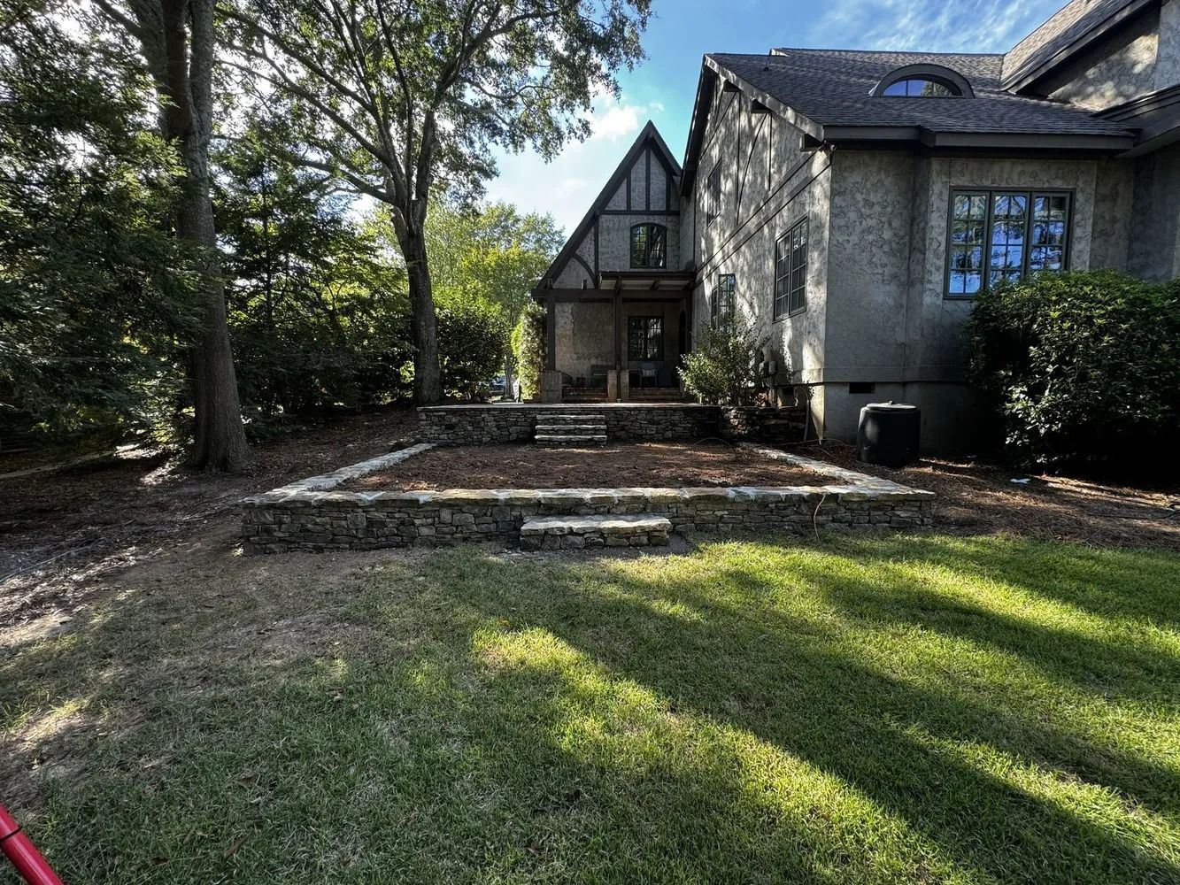 Front yard of a stone house with a brick walkway, steps, and long tree shadows across the lawn
