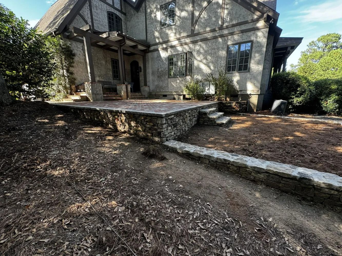 Front of a stone house with a raised patio and dirt yard, seen on a sunny day.