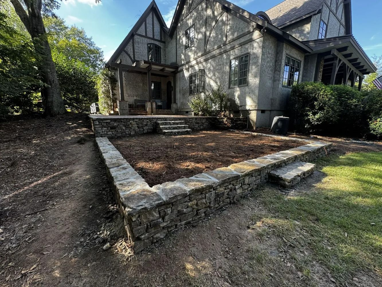 Stone-front Tudor-style house with a raised front garden bed and steps, surrounded by trees and shrubs