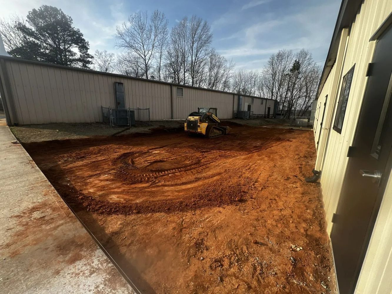 Small construction site with a skid steer leveling red dirt beside a building and long fence.