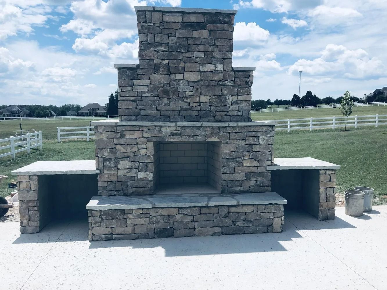 Stone outdoor fireplace with side benches on a concrete pad in a grassy field