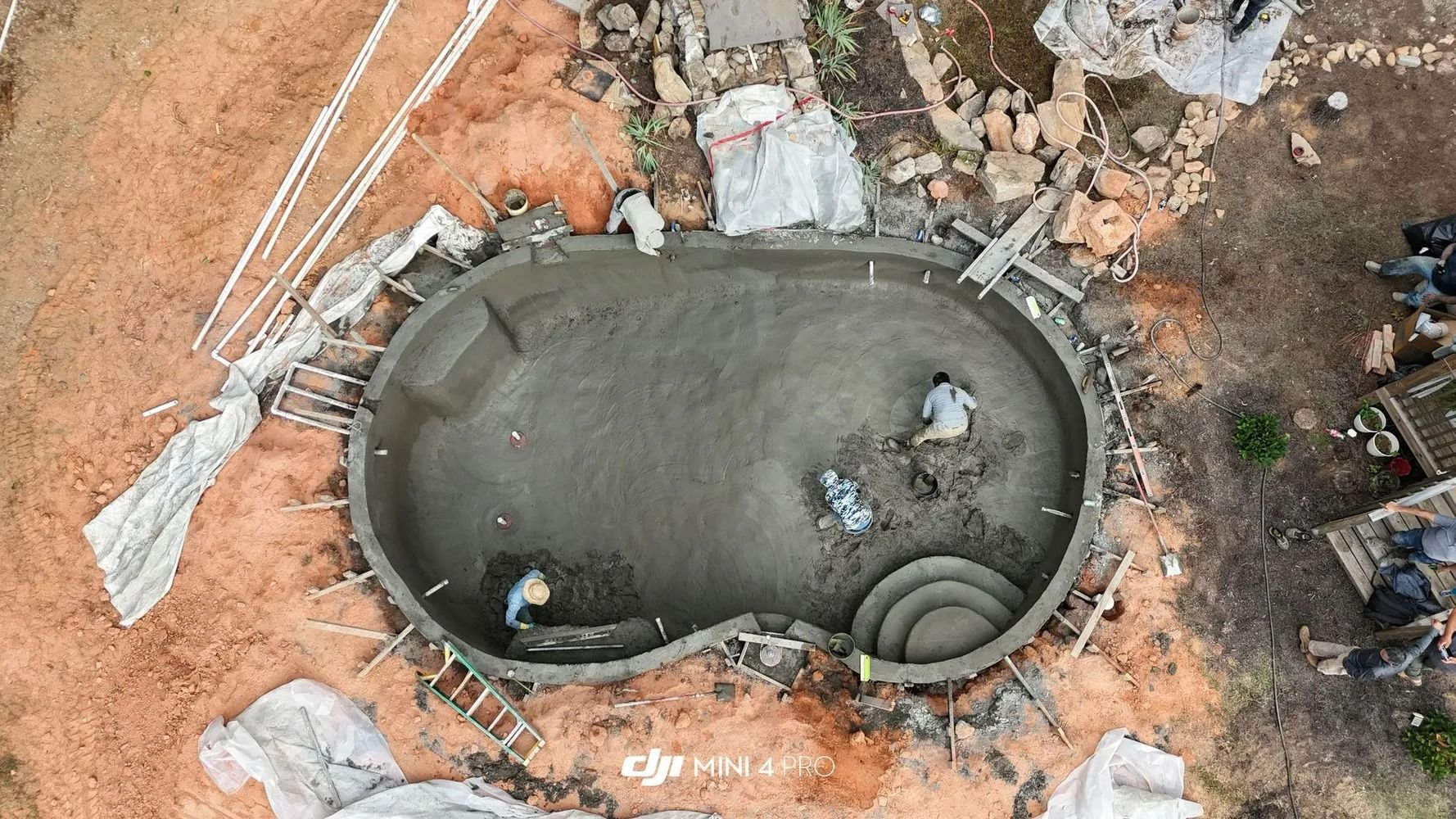Aerial view of a small oval concrete pool under construction, with workers and coiled hoses around it.