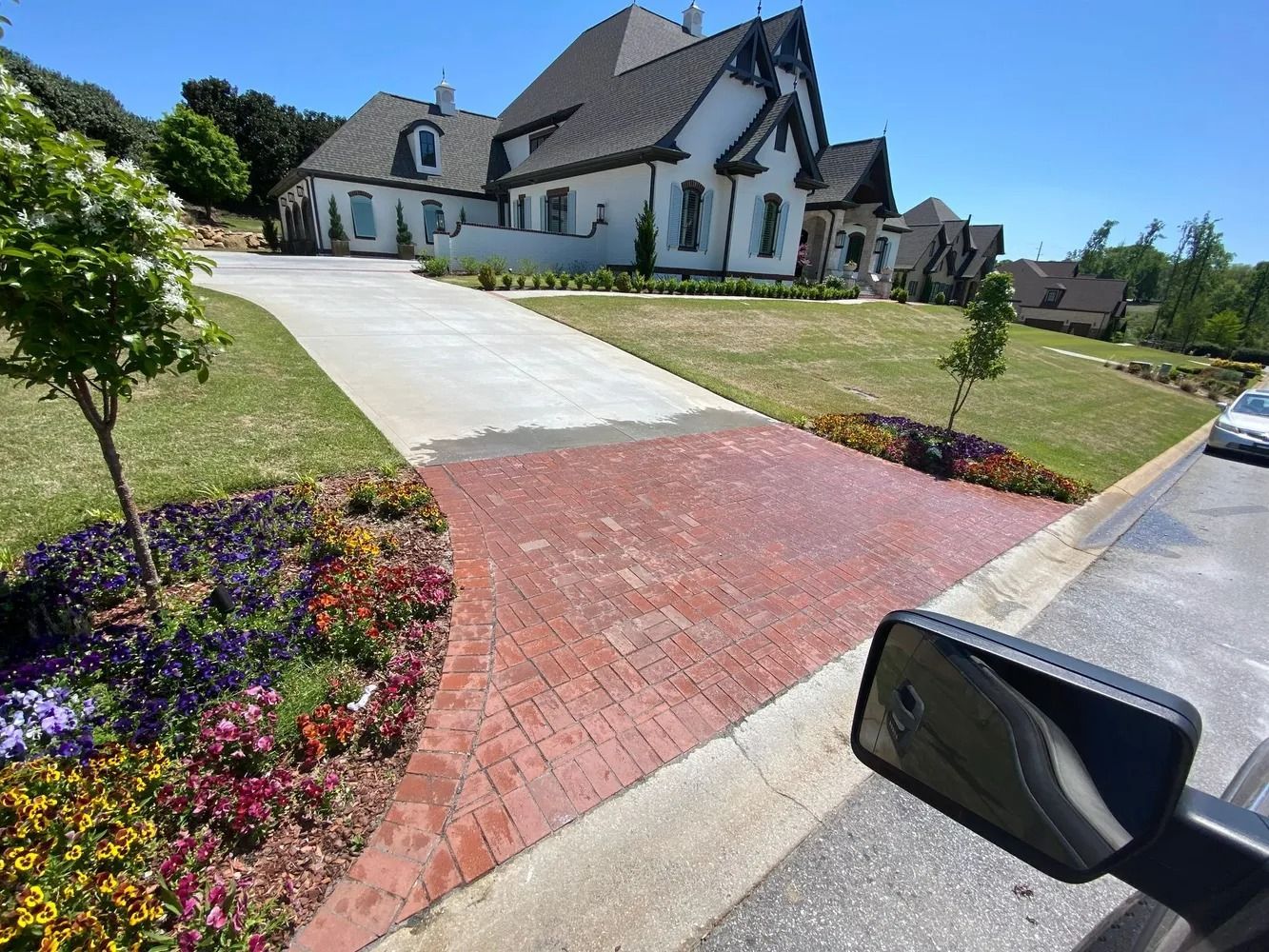 Large white house with a steep roof, red brick driveway, and landscaped flower beds on a sunny day