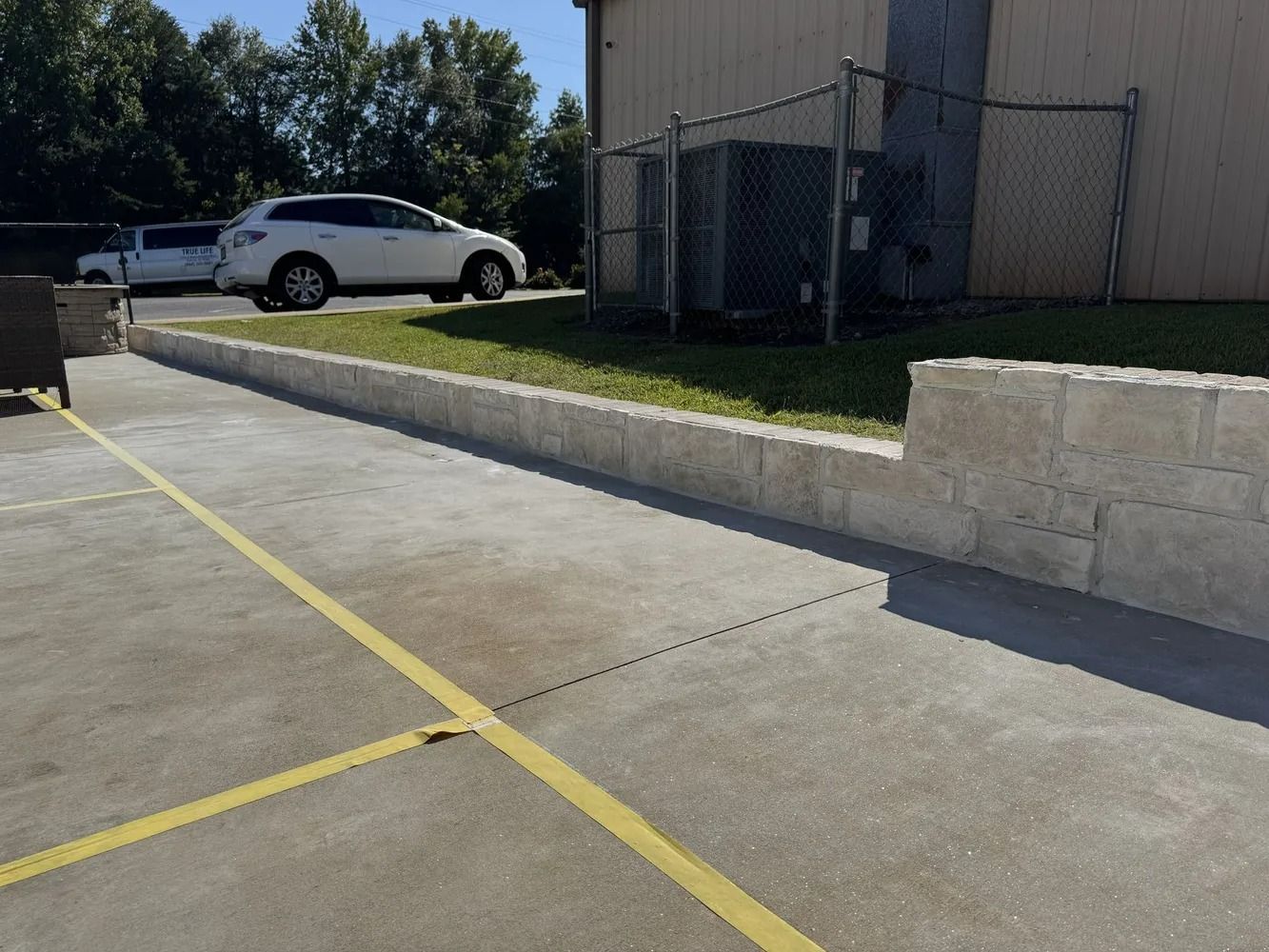 Concrete parking lot with yellow lines beside a white SUV and a building wall under clear sunlight