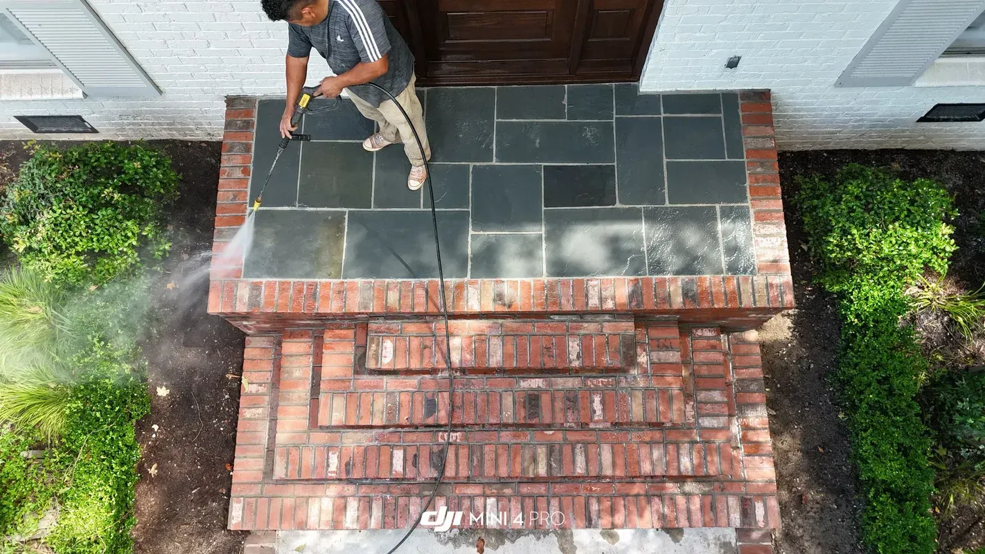 Worker pressure-washing a dark roof next to red brick siding and green shrubs