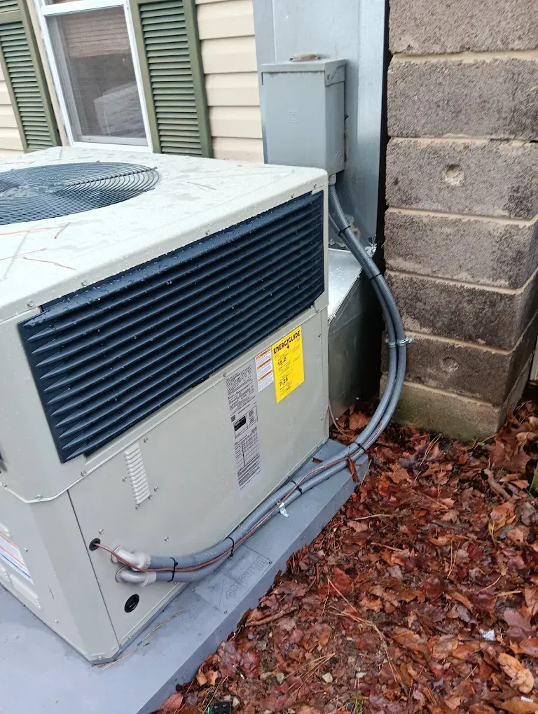 Outdoor air conditioner unit beside a house wall, with hoses and fallen leaves on the ground.