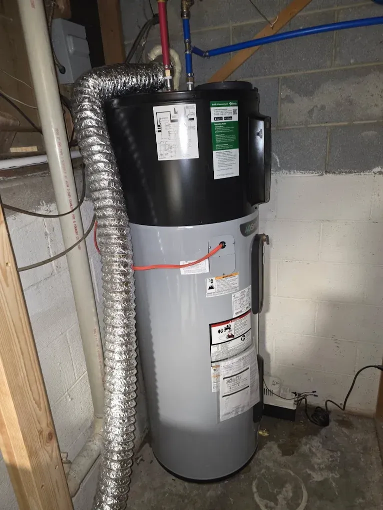 Water heater in a basement, with black and gray tank, vent pipe, and utility labels.