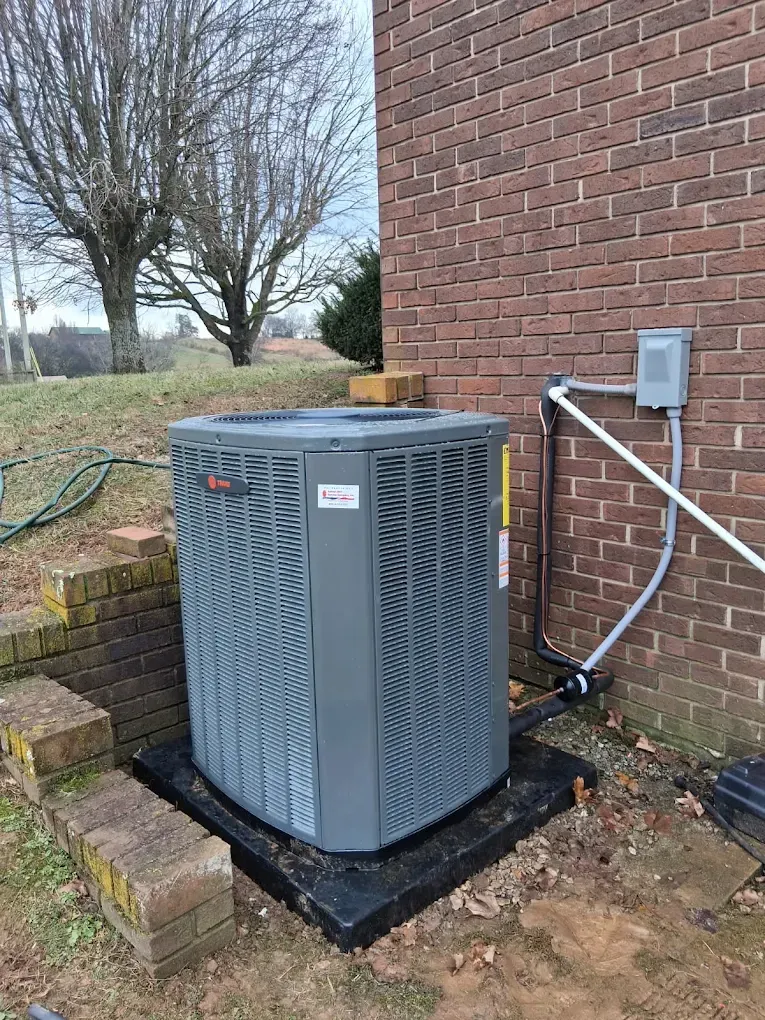 Outdoor HVAC unit beside a brick house with conduit and piping on a concrete pad