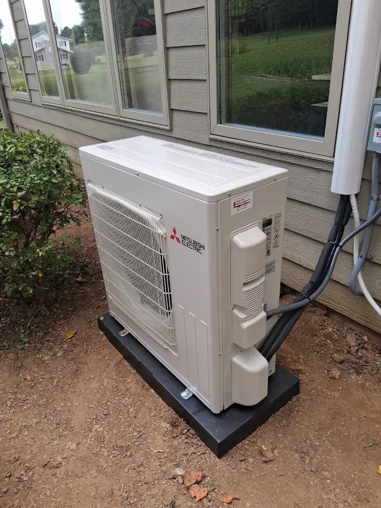 Outdoor HVAC unit beside a building, with white casing and black base on gravel ground
