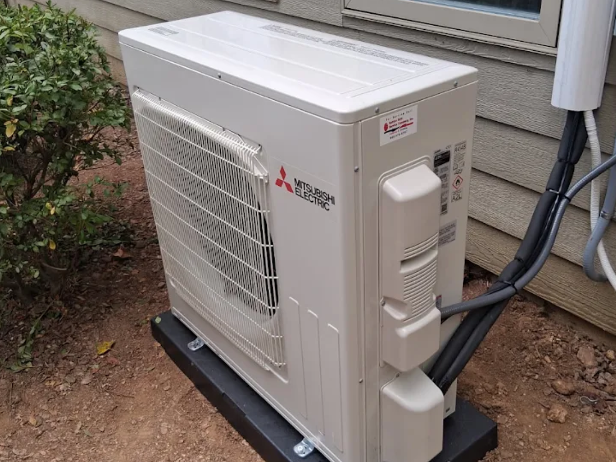 Outdoor Mitsubishi mini-split AC unit beside a house on a metal stand, with pipes and conduit attached