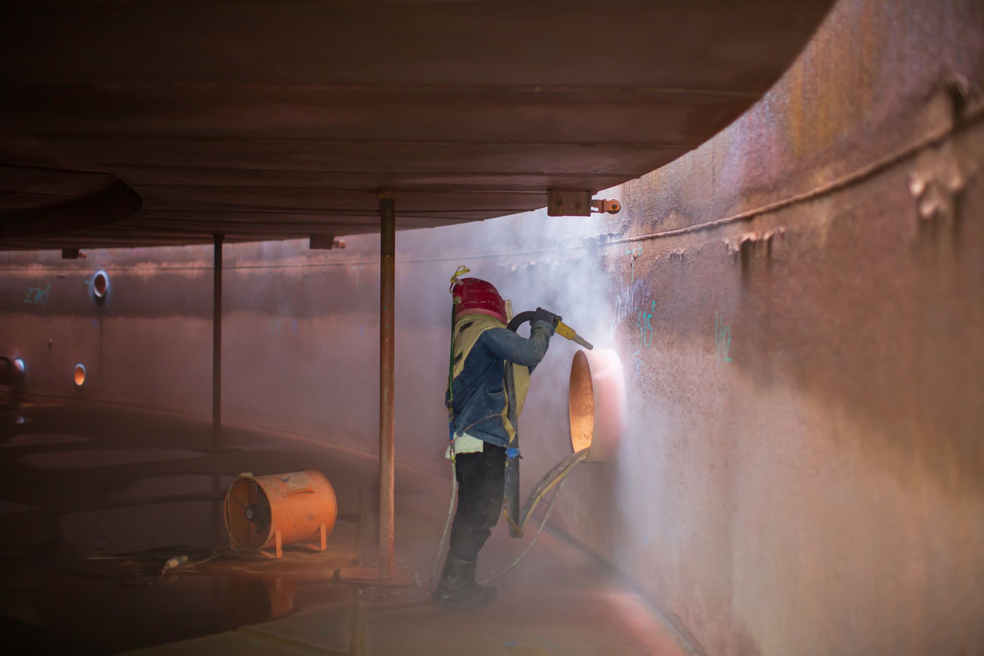 A worker is sandblasting a surface for corrosion plate preparation of an internal oil tank.