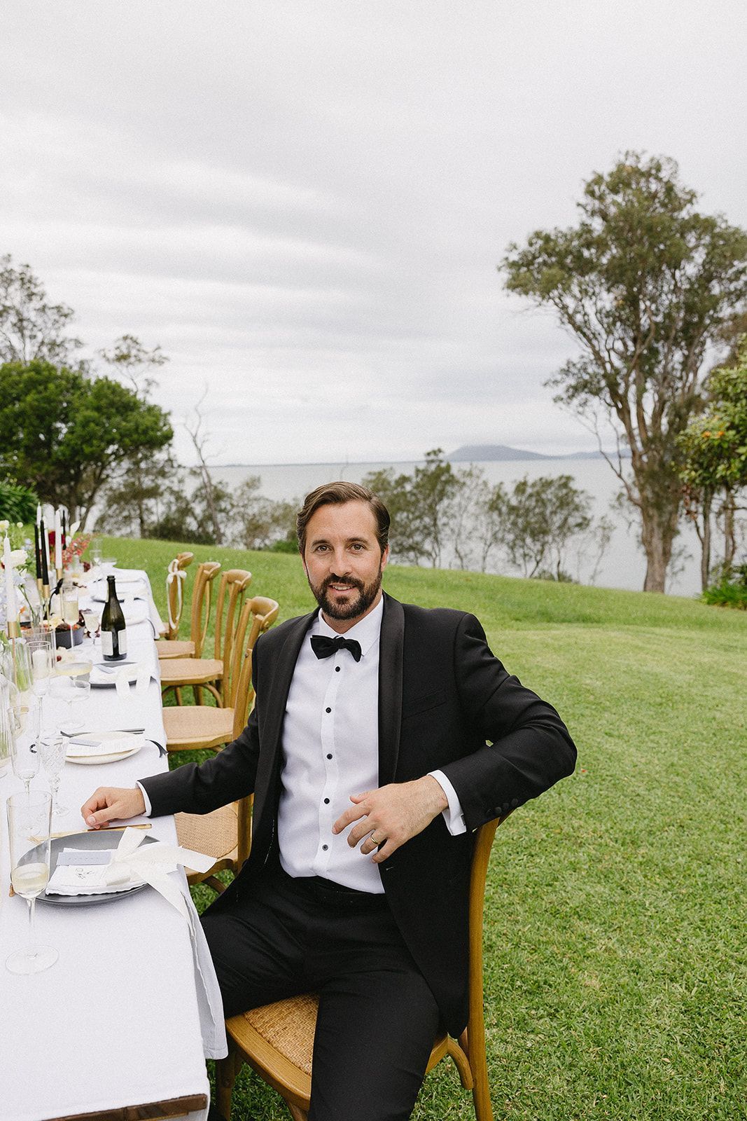 A man in a tuxedo is sitting at a long table in a field.