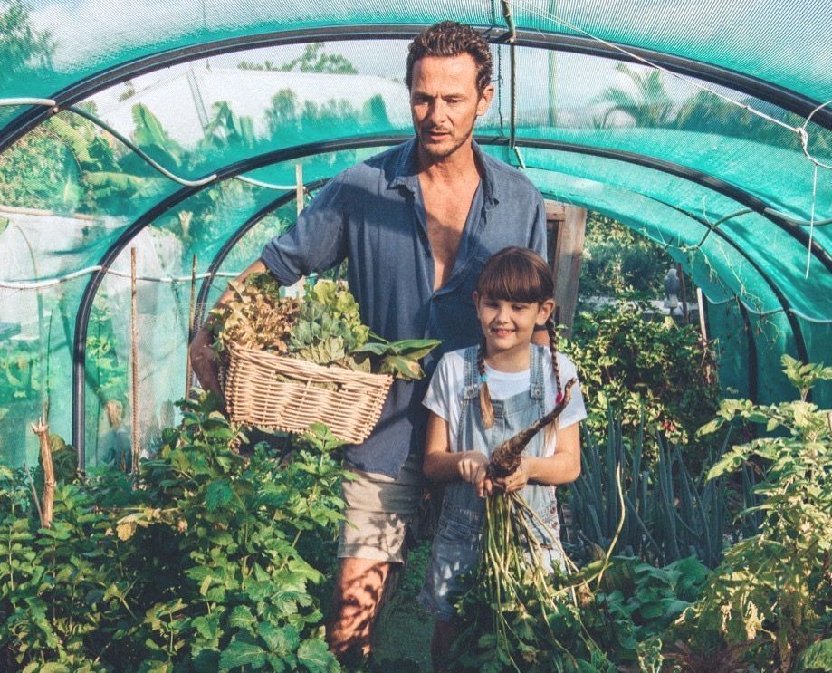 A man and a little girl are standing in a greenhouse holding baskets of vegetables.