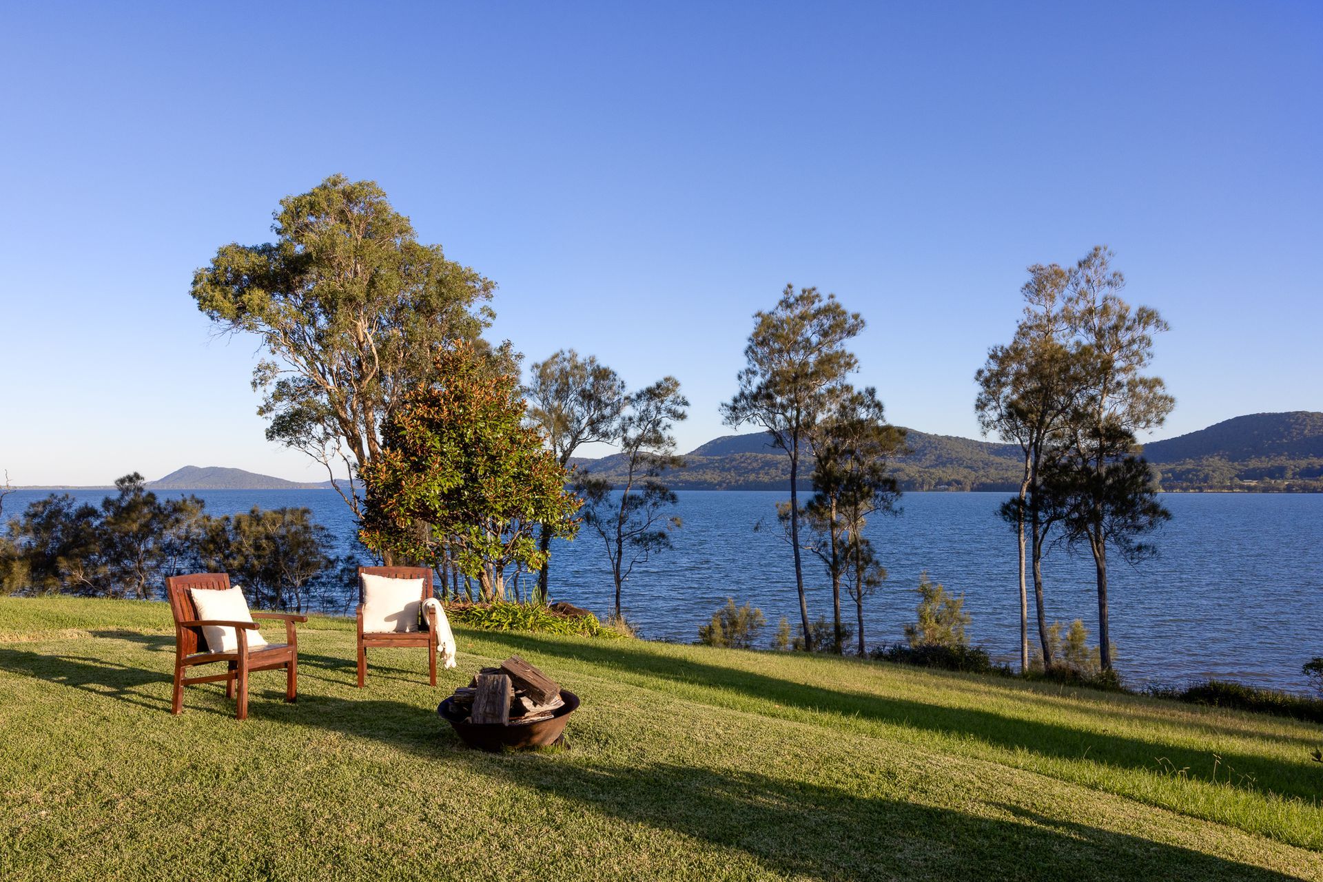 Two chairs and a fire pit on a lush green lawn overlooking a lake.