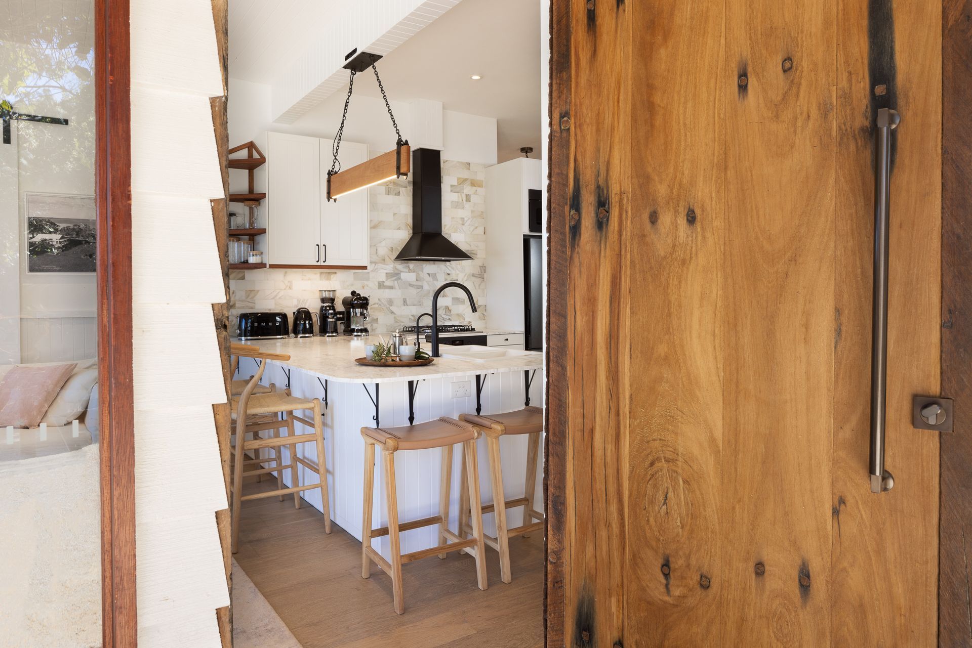 A wooden door is open to a kitchen with white cabinets and stools.