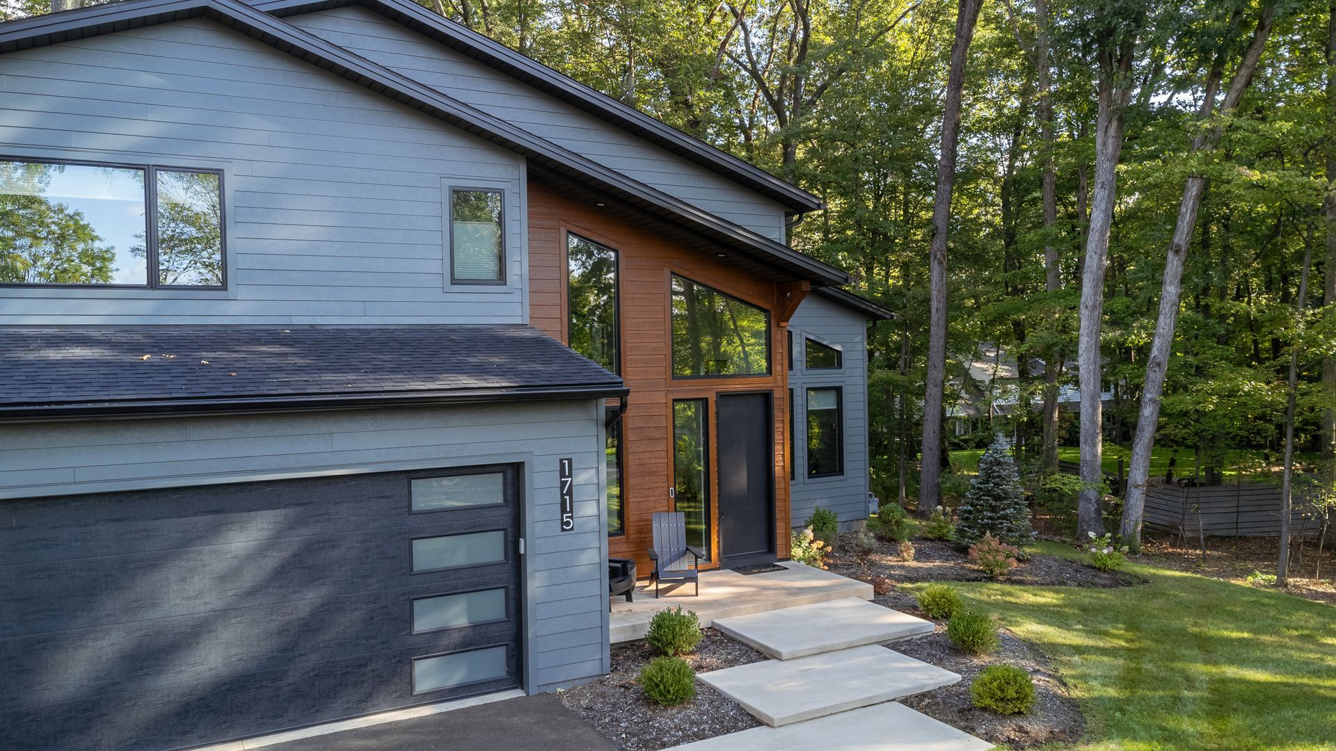 Modern house exterior with gray garage, stone walkway, large windows, and wooded yard