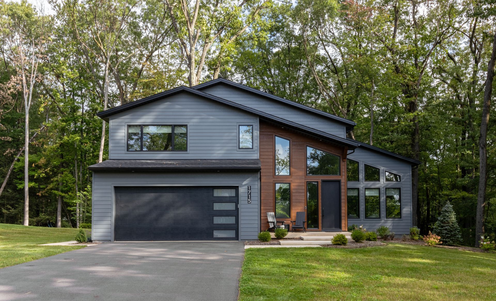 Modern two-story house with dark gray siding, brick accents, and a garage surrounded by trees