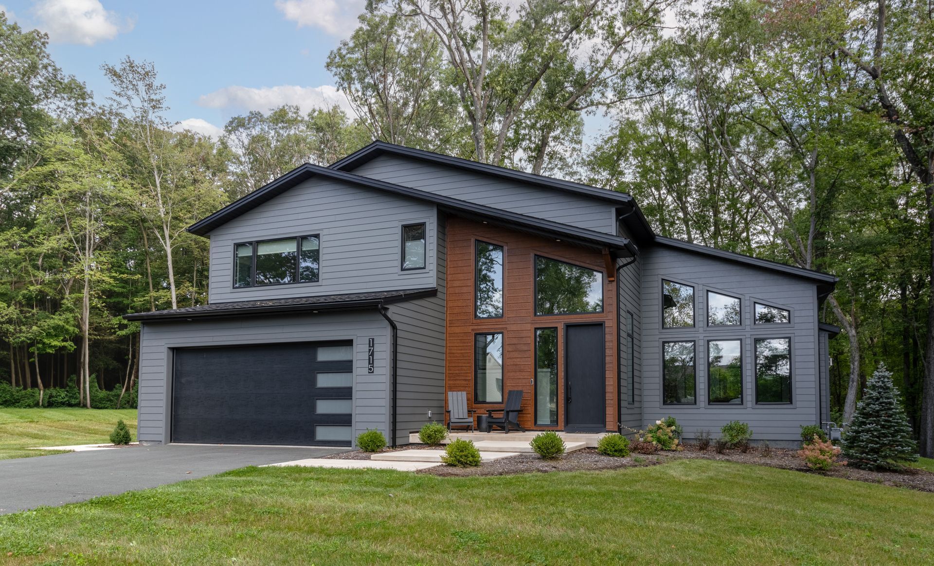 Modern two-story gray house with black trim, large windows, and a garage, surrounded by trees and lawn