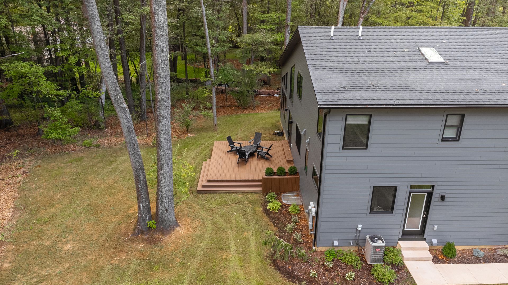 A gray house with a wooden deck and patio furniture, surrounded by trees and a grassy yard.