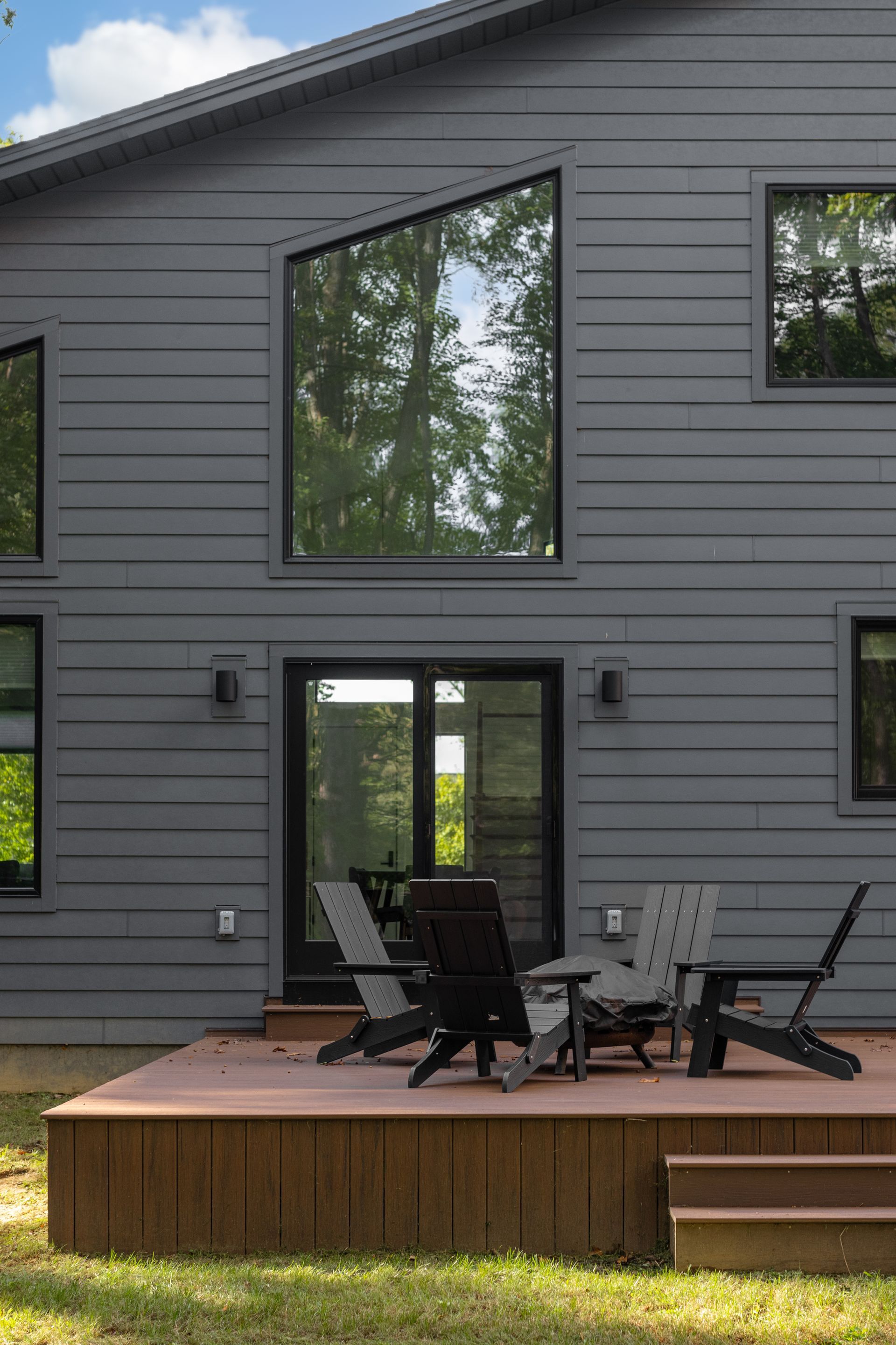 Gray house exterior with large windows and a wooden deck furnished with black patio chairs and table
