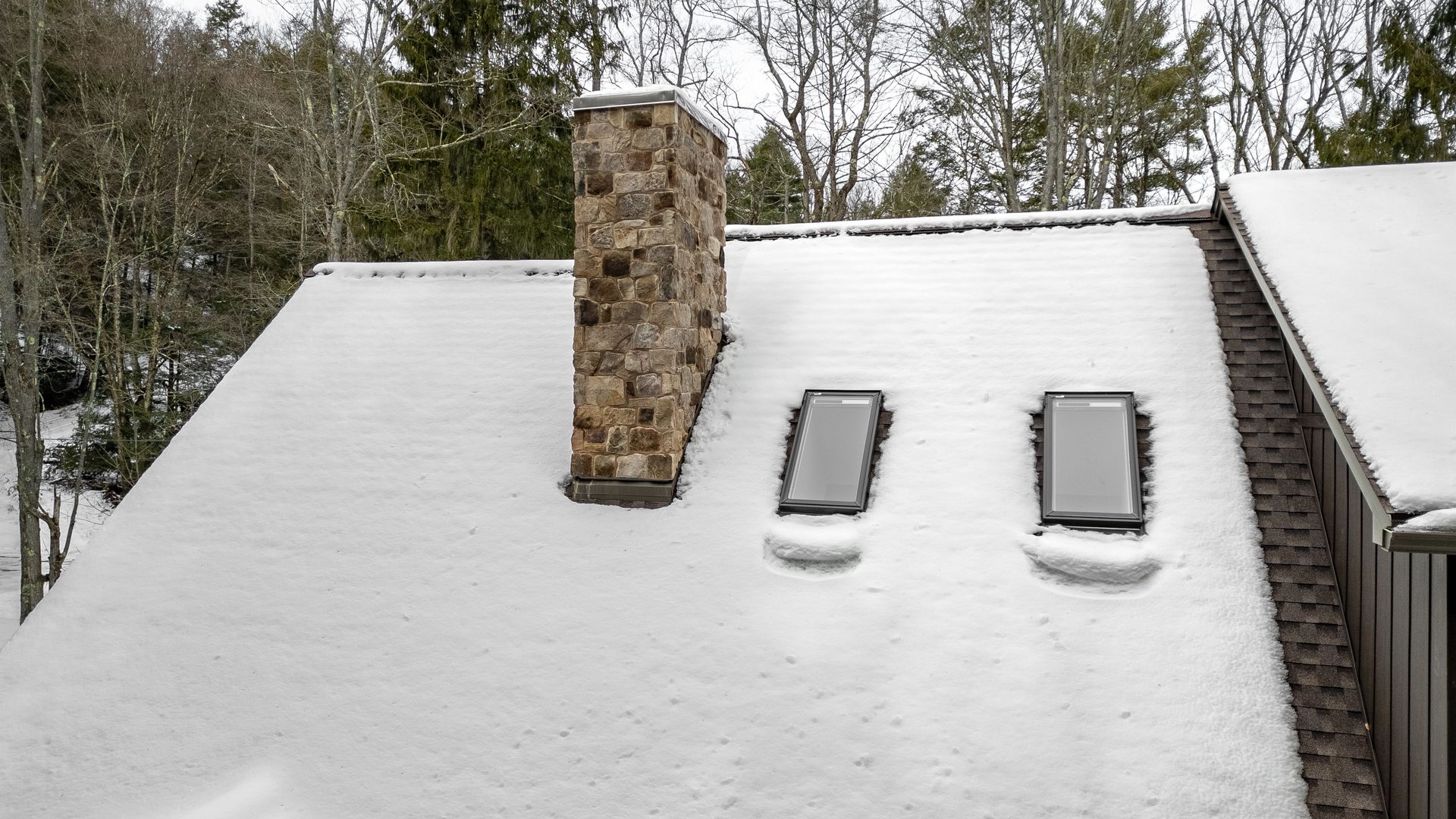 Snow-covered rooftop with two skylights and a stone chimney beside trees