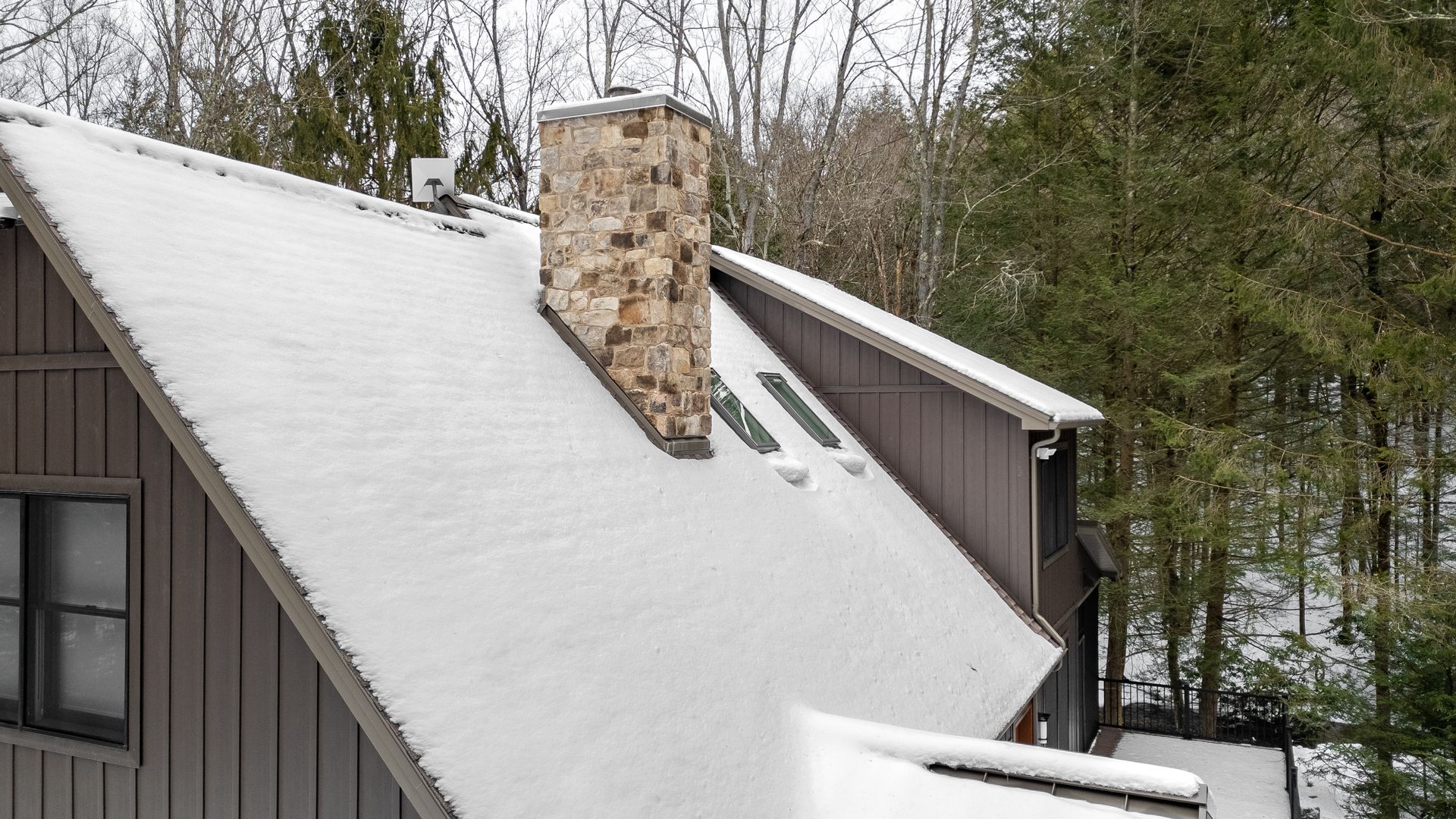 Snow-covered house roof with a stone chimney and trees in the background